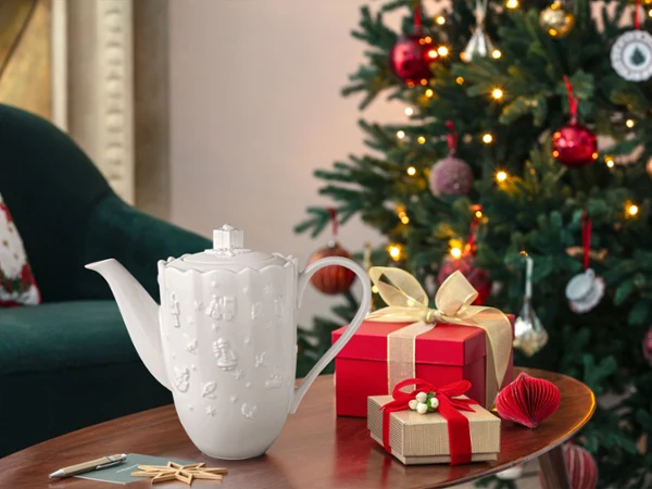 Two wrapped gifts and a white Toys Delight Royal Classic ceramic jug stand on a wooden table next to a decorated Christmas tree.