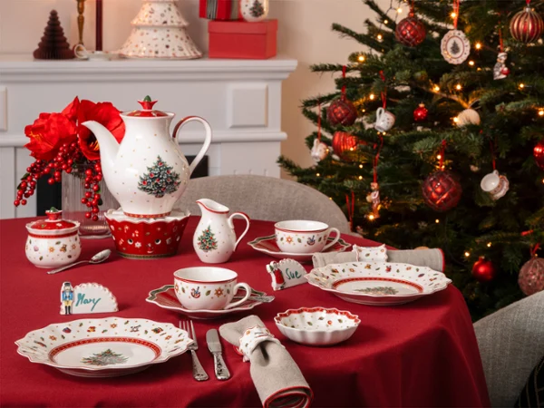 A table set with crockery and cutlery A table set with crockery and cutlery featuring Christmas motifs, next to a decorated Christmas tree and an illuminated mantelpiece.