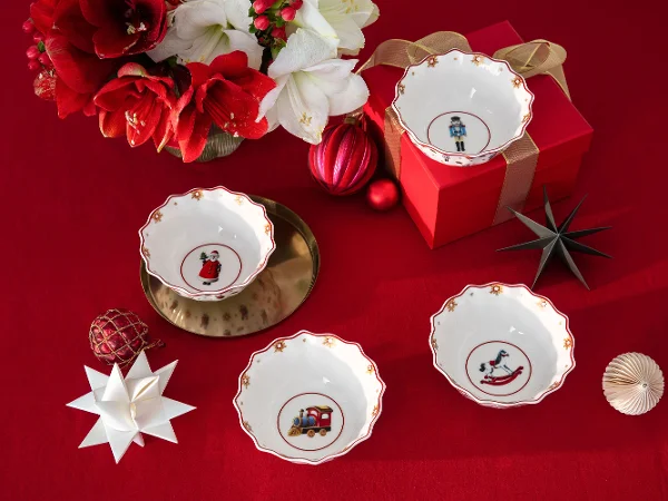 Quattro ciotole decorative con motivi natalizi sono presentate su una tovaglia rossa con ornamenti festivi e una confezione regalo.