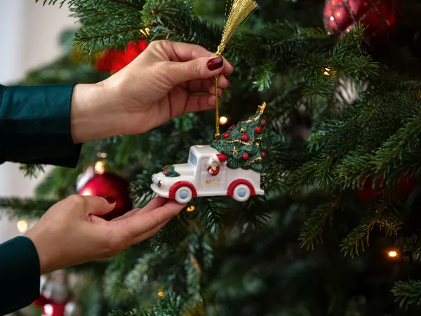 Un ornement en forme de camion sur un sapin de Noël décoré Une personne accroche un petit ornement Villeroy & Boch en forme de camion sur un sapin de Noël décoré, orné de boules rouges et de lumières.