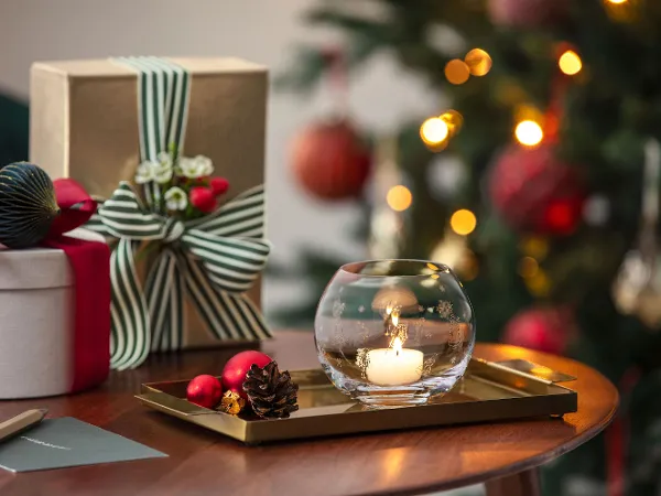 Une bougie allumée dans un porte-bougie à réchaud Toy's Delight Une bougie allumée dans un porte-bougie à réchaud Toy's Delight est posée sur un plateau à côté de cadeaux emballés, avec un sapin de Noël décoré en arrière-plan.