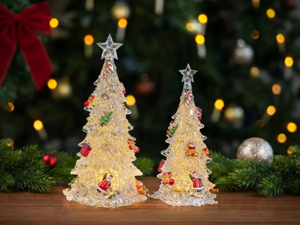 Two Christmas tree lamps stand on a wooden base; a Christmas tree and Christmas decorations can be seen in the background.