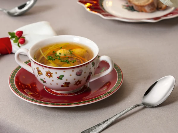 Toys Delight Soup Cup by Villeroy & Boch. A cup of clear soup with herbs sits on a decorated saucer next to a spoon on a neatly set table.