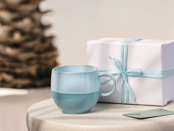 Hellblaue Perlemor Aqua Tasse und weiße Geschenkbox mit blauem Band auf einem runden Tisch; unscharfer Hintergrund mit einer Pflanze.
