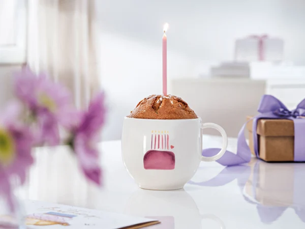 Na stole babeczka z filiżanki ze świeczką w kubku With Love Birthday, obok fioletowe kwiaty i beżowe pudełko prezentowe.