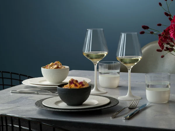 Elegant dinner with two white wine glasses, neatly arranged plates and serving bowls from Manufacture with dishes on a modern table.