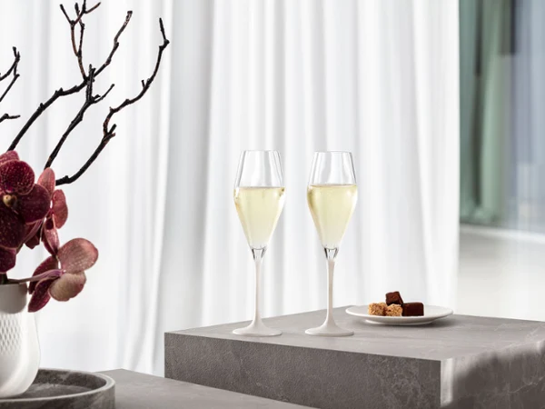 Dwa kieliszki do szampana Manufacture z białą nóżką na szarej powierzchni Dwa kieliszki do szampana Manufacture z białą nóżką na szarej powierzchni, obok talerza z pralinkami i wazonu z różowymi kwiatami i gałązkami.