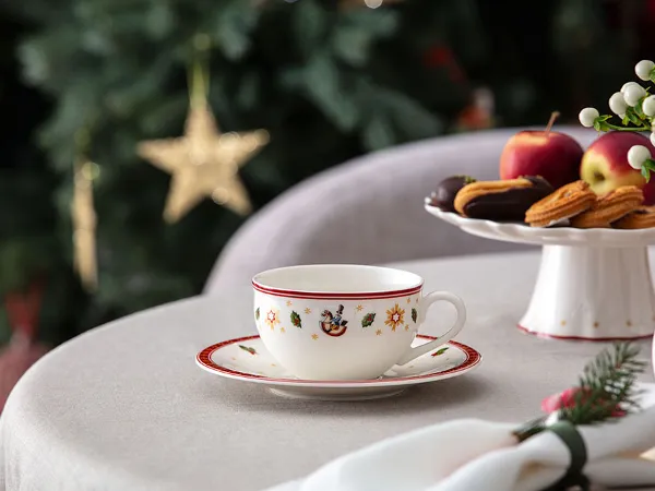 Une tasse à thé et une sous-tasse « Toy's Delight » de Villeroy & Boch Une tasse à thé et une sous-tasse « Toy’s Delight » de Villeroy & Boch sont posées sur une table à côté d’une assiette contenant des biscuits et des pommes, avec un sapin de Noël décoré en arrière-plan.