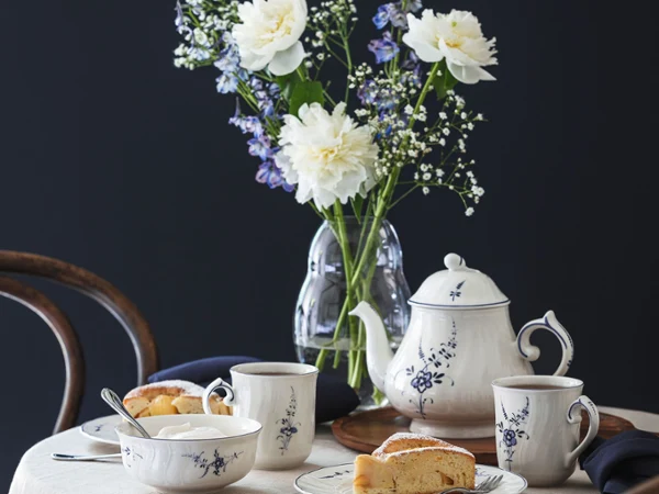 A table covered with a white tablecloth for afternoon tea, with an Old Luxembourg floral-patterned teapot, cups, plates with slices of cake and a vase of white and purple flowers against a dark background.
