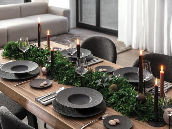 Elegant dining table set with black Manufacture plates, black candles, cutlery and green foliage as a centrepiece in a modern dining room.