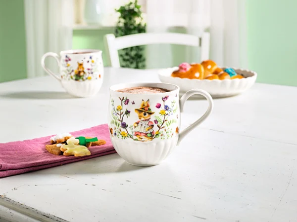 Dos tazas de Pascua sobre una mesa con servilleta rosa y flor, al fondo un cuenco de Pascua con pan de Pascua