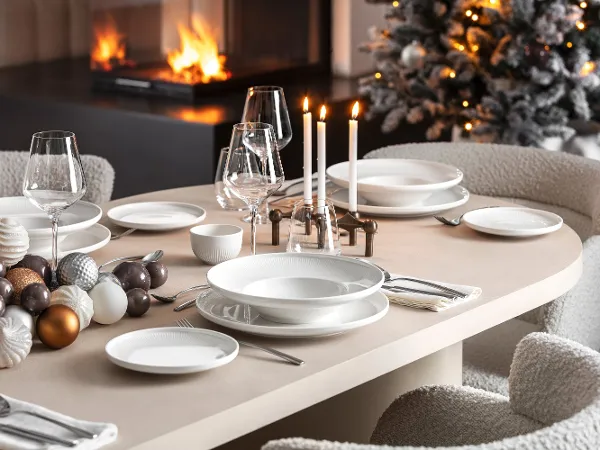 Festive dining table with white Afina crockery A festive dining table with white Afina crockery, candles and decorative ornaments from Villeroy & Boch, next to a burning fireplace and a decorated Christmas tree in the background.