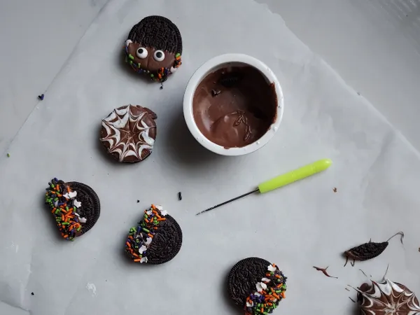 Halloween cookies with sprinkles and a spider web pattern, next to melted chocolate and a green knife.