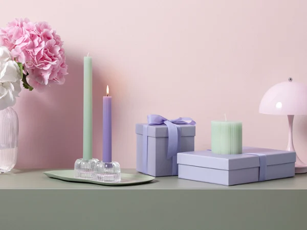 Velas en tonos verde pastel y violeta de Fleur como idea para regalo Velas largas en tonos pastel de Fleur, cajas de regalo y flores dispuestas sobre una superficie verde frente a una pared rosa pálido con una pequeña lámpara.