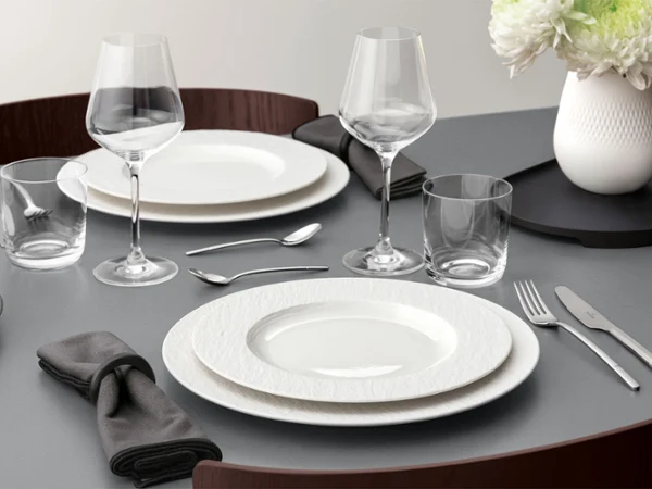 A formal table setting with a white plate on a white plate, glassware, and silver cutlery on a grey table.