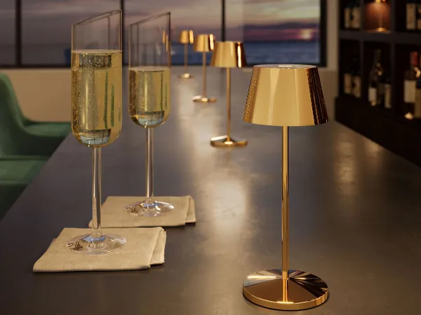 Seoul Leuchte in Gold neben 2 Champagnergläsern Zwei Champagnergläser auf Servietten und eine goldene Leuchte stehen auf einer dunklen Bar mit Regalen und einem Blick auf den Sonnenuntergang im Hintergrund.