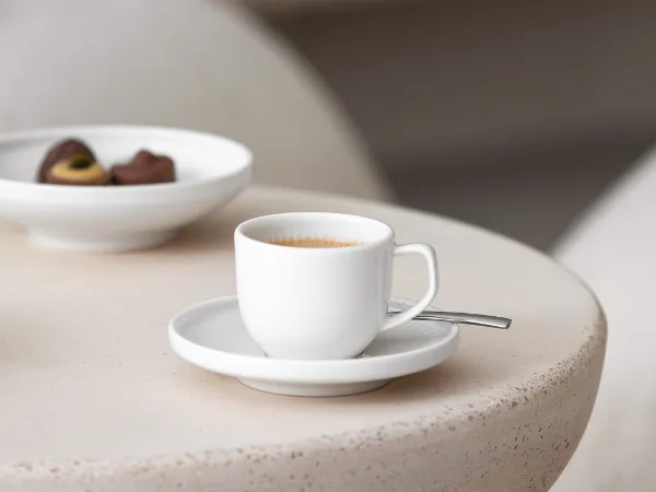 Une tasse à expresso avec sa sous-tasse Afina de Villeroy & Boch avec des chocolats et une cheminée allumée en arrière-plan.