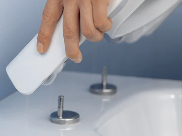 Fonction QucikRelease pour un couvercle de WC Gros plan d’une main tenant, sur un fond bleu, une buse de bidet au-dessus d’une robinetterie de salle de bains blanc à deux commandes.