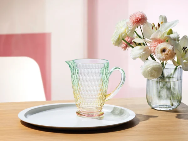 On a wooden table, a Boston glass jug stands on a white tray next to a vase of white and pink flowers.