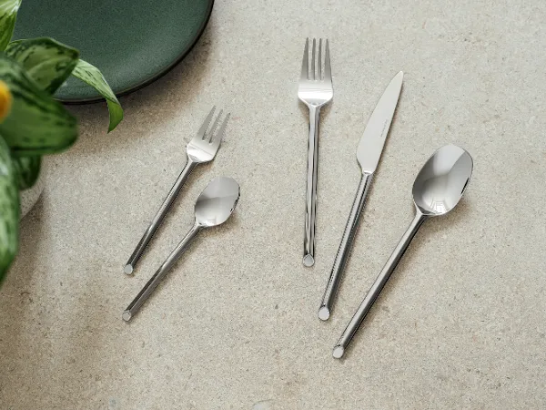 Five like cutlery pieces from like.by Villeroy & Boch- fork, knife, spoon, dessert fork and teaspoon Five pieces of cutlery in Villeroy & Boch style - fork, knife, spoon, dessert fork and teaspoon - are arranged on a light-coloured stone surface next to a green plate and a plant.