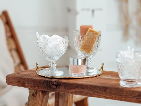 A golden tray with two Boston glasses on a wooden stool A golden tray with two Boston jars containing cotton balls and soap as well as a small jar of cream on a wooden stool.