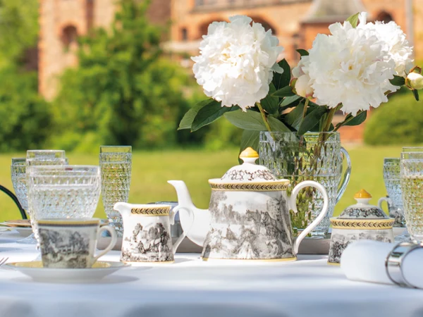 A table set for outdoor tea with ornate teacups and an Audun teapot, crystal glasses, and white peonies in a vase. A historic building is blurred in the background.