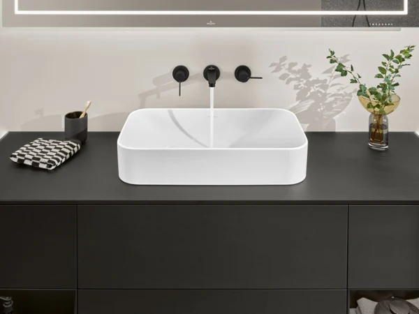 Finion Waschbecken mit schwarzer Armatur und Arbeitsplatte Ein stilvolles Villeroy & Boch Waschbecken mit einem weißen rechteckigen Becken, einer schwarzen Arbeitsplatte, einem schwarzen Wasserhahn, einem gefalteten Handtuch, einem Zahnbürstenhalter und einer Vase