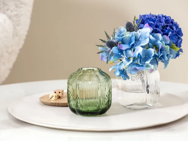 „Fleur Vasen“ von Villeroy & Boch, eine kleine grüne und durchsichtige Vase mit blauen Blumen. „Fleur Vasen“ von Villeroy & Boch, eine kleine grüne und durchsichtige Vase mit blauen Blumen, steht auf einem runden weißen Tablett mit Holzwürfeln auf einem Glastisch.