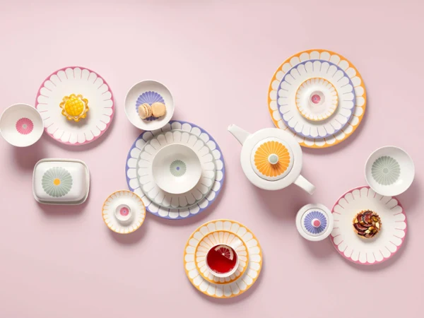 Various patterned ceramic bowls, dishes, and a teapot from the Fleur collection on a pink background, several plates with colorful desserts.