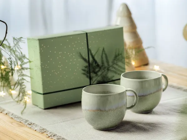 Green Perlemor cups with gift packaging Two green Perlemor cups on a table with a matching green gift box in the background, decorated with a tree and lights.