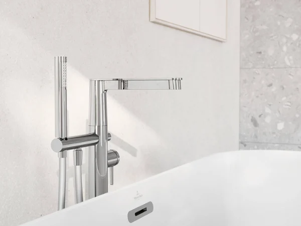 Una moderna bañera cromada con mango de ducha instalada delante de una pared de azulejos blancos. La bañera blanca es parcialmente visible en primer plano.