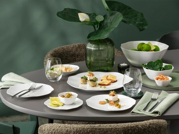 A round dining table set with white tableware from the Pura collection A round dining table set with white tableware from the Pura collection, including starters, glasses, napkins and a centrepiece of green plants.