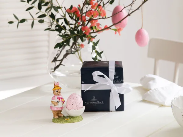 A Villeroy & Boch Bunny Tales egg cup on a white table next to a gift box wrapped with a bow. In the background is a vase with twigs and pink flowers.