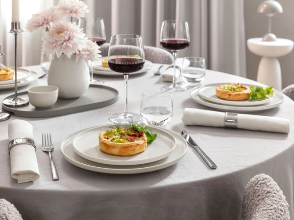 Mesa elegante para cuatro personas con vajilla de porcelana Afina y copas de vino tinto Maxima llenas de vino. Servida con comida en un ambiente moderno y refinado.