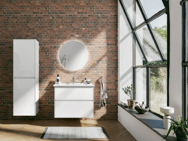 Les collections de salles de bains Collaro et Venticello dans une salle de bains de type loft aérée Une salle de bains loft moderne avec des collections de salle de bains de Villeroy & Boch, un mur de briques, une grande fenêtre, un lavabo blanc avec un miroir rond, un tapis et une plante en pot.