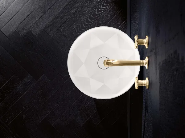 Octagon Waschbecken mit goldener Armatur Draufsicht auf ein weißes, facettiertes Waschbecken mit goldener Armatur und Griffen, an einer schwarzen Wand montiert. Der Boden besteht aus dunklem Fischgrätholz.