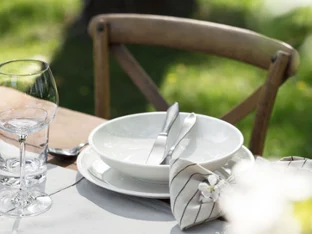 Artesano plate outdoors on a wooden table Outdoor dining area with an empty white Artesano plate, flatware, and a glass on a wooden table.
