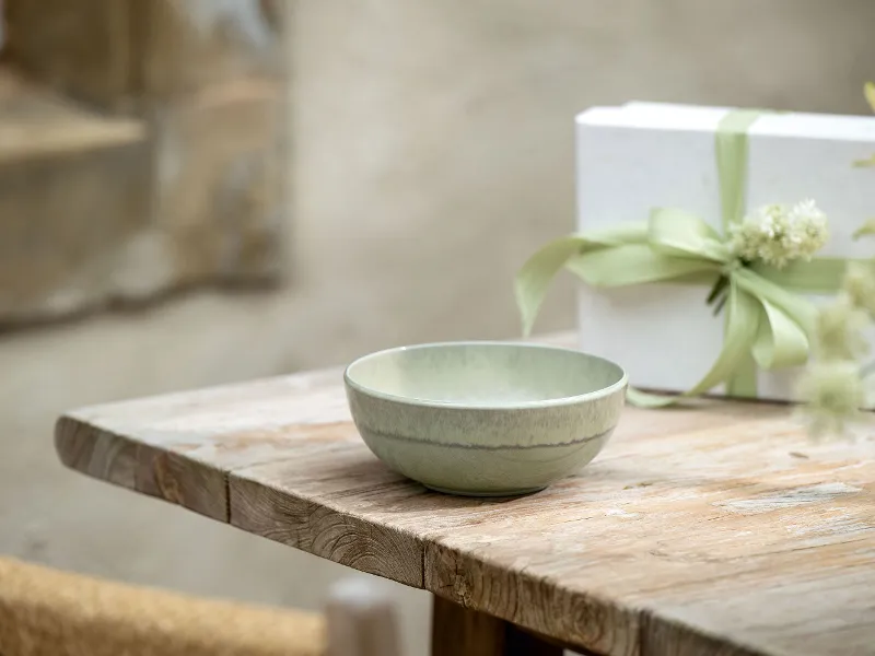 On a wooden table, there is a ceramic bowl next to a white gift box tied with a green ribbon.