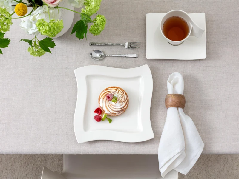 Dessert on a square NewWave plate with raspberries, napkin, spoon, fork, and a cup of tea on a table with a light tablecloth and floral decoration.