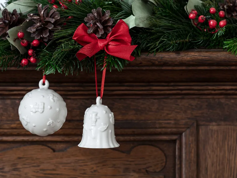 Les ornements en forme de boule et de cloche blanches Toy's Delight Royal Classic de Villeroy & Boch sont suspendus par des rubans rouges à une guirlande décorée avec des pommes de pin et des baies rouges, ornant une surface en bois.