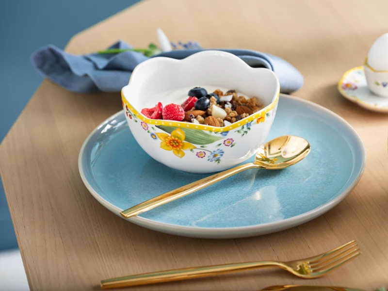 A breakfast set consisting of a Spring Awakening cereal bowl and a light-blue Crafted plate, with a gold spoon and a cup of tea on the side.