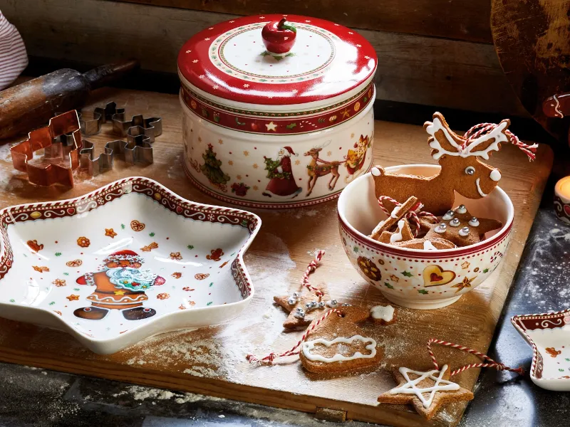 Świątecznie nakryty drewniany stół z ceramicznymi naczyniami „Winter Bakery Delight” marki Villeroy & Boch oraz porozrzucane ciasteczka, ozdobne blaszane pudełko, foremki do ciastek i mąka do pieczenia.