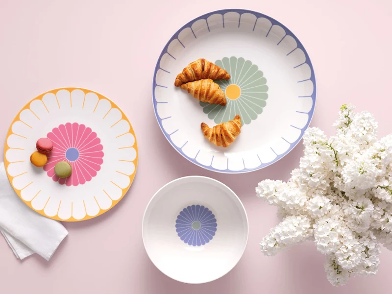 Three Villeroy & Boch Fleur plates with croissants and macarons next to white flowers on a light pink background.