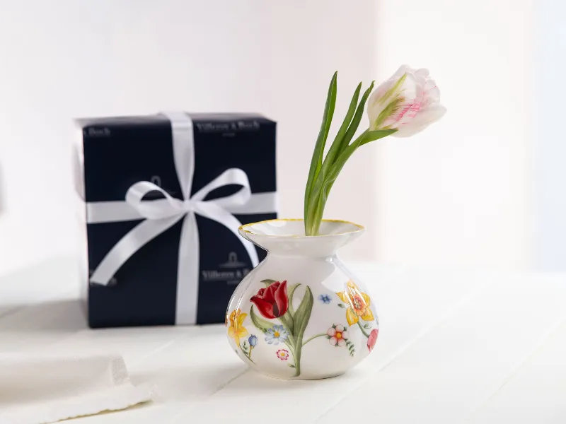 A Spring Awakening flower vase with two tulips next to a gift box wrapped in a white bow on a white table.