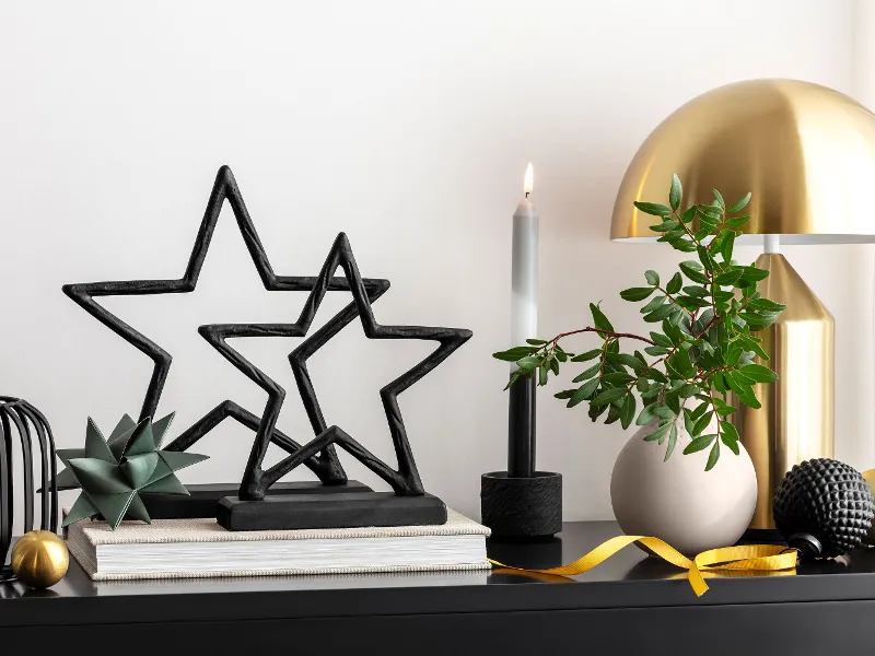 Une décoration de table avec des sculptures d’étoiles noires Black Xmas, une lampe dorée, des livres, une bougie allumée et divers objets décoratifs.