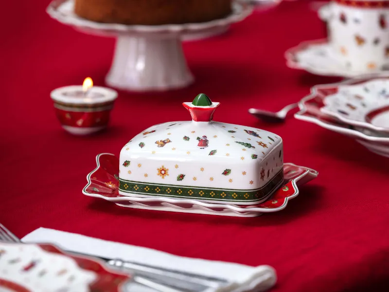 Toy's Delight butter dish with a festive pattern on a red tablecloth, surrounded by festive plates and an unlit candle in the background.