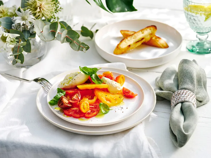 The Crafted Cotton dinner set for two, featuring fresh tomatoes and bread, is complemented by green Boston glassware, green linen napkin, and accented with a vase of florals. Set on a white linen cloth atop a white wooden table.