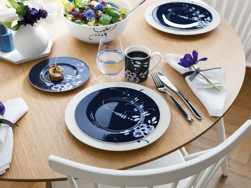 A round wooden table with blue and white plates, cups and a small dessert. In the centre is a salad bowl and blue flowers.