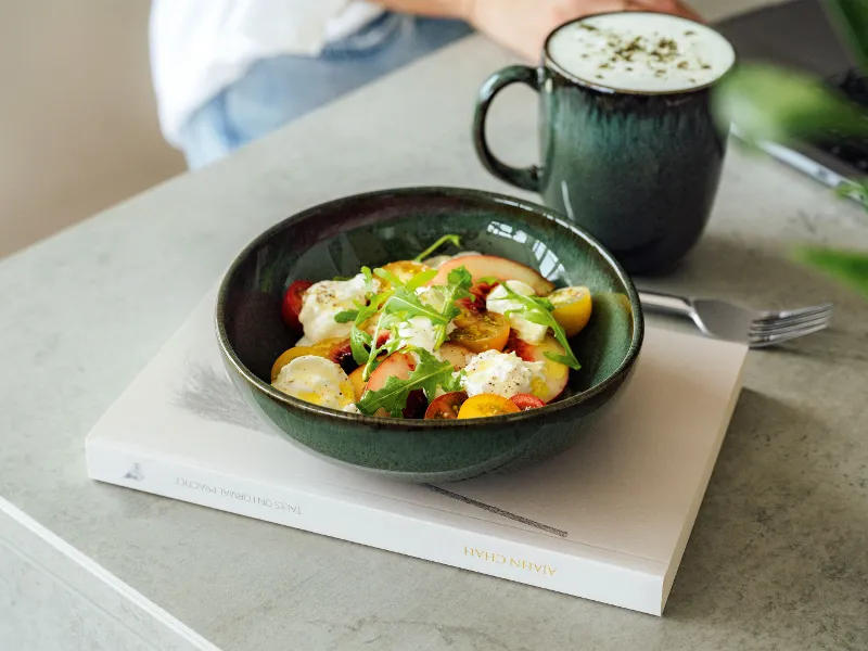 A salad with tomatoes, lettuce and cheese is presented on a book in a green Lave bowl from Villeroy & Boch, accompanied by a frothy drink in a matching green mug on a grey background.
