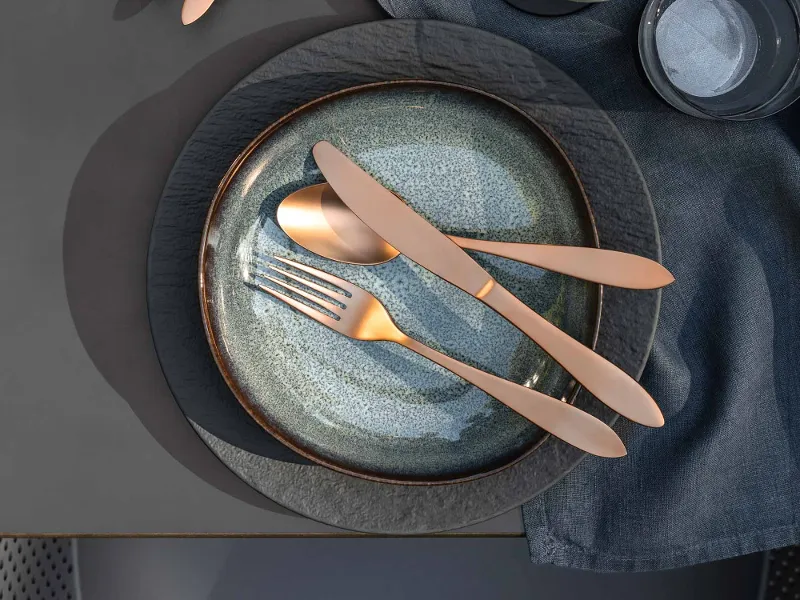 Elegante Tischdekoration mit zwei Lave Tellern, Manufacture Besteck, einer Schüssel Oliven und einem Glas Weißwein auf einer grauen Tischdecke.